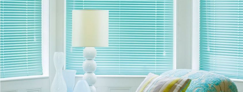 Teal Venetian blinds in a bright bay window beside a decorative lamp and patterned cushions.